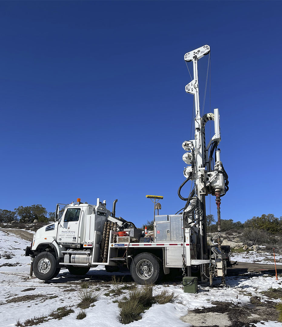 A truck-mounted drill rig with its mast raised on frost-covered ground under a clear sky