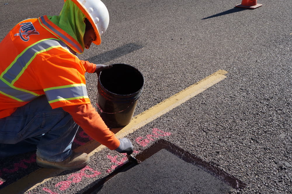 A crew member in high-visibility gear finishing the edge of a fresh asphalt patch in the road.