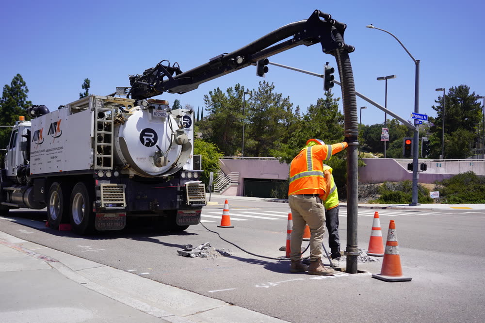 A vacuum excavation truck with its boom extended over a coned-off pothole, an operator guiding the hose.