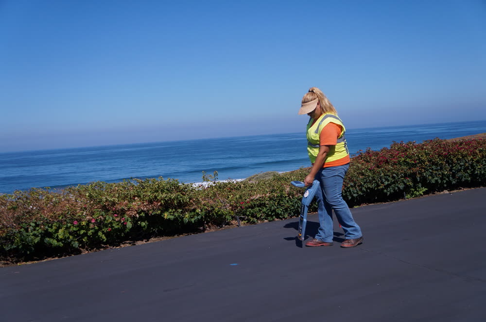 A locator in high-visibility gear sweeping a sidewalk with a utility locating receiver, the ocean behind.
