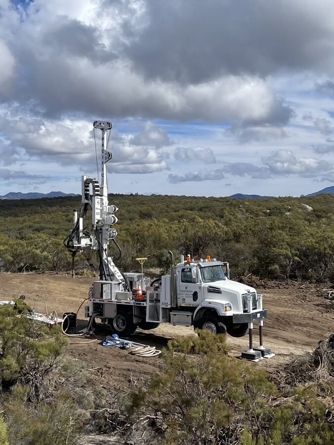 A white truck-mounted drill rig set up on an open hillside site.