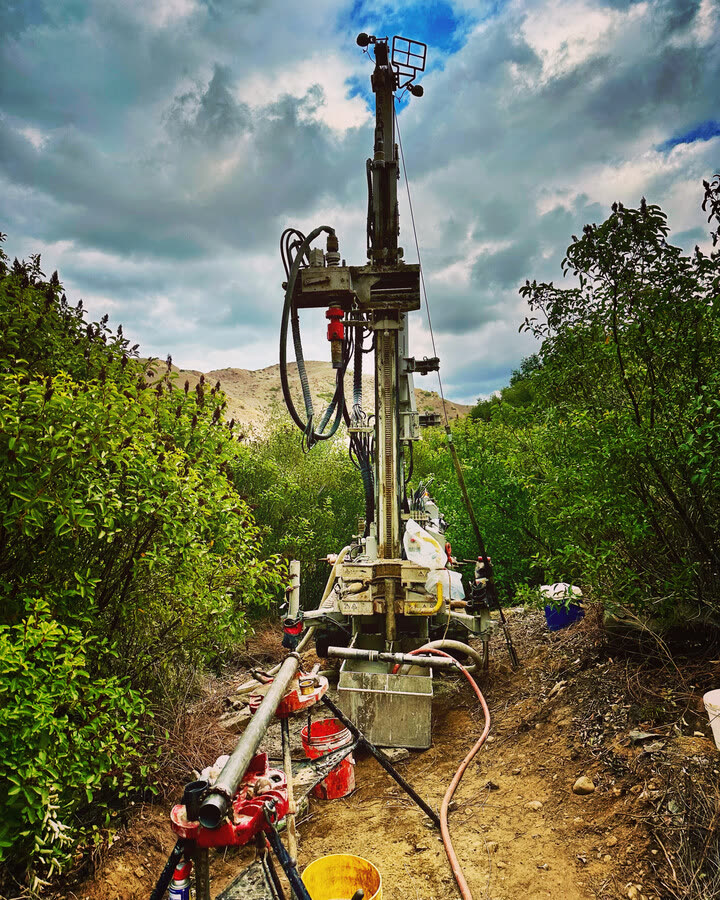 A compact track-mounted rig drilling on a steep, brush-covered slope.