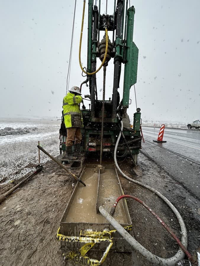 A driller in high-visibility gear working a rig on wet, muddy ground.